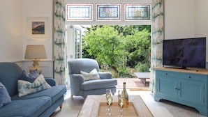 A living room with a blue sofa, a wooden coffee table with glasses and a bottle, a television on a blue cabinet, and a view of greenery through a glass door. - Fern Bank (Penrith)