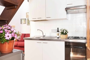 Une cuisine moderne avec des armoires blanches, un évier en acier inoxydable, une cuisinière à gaz et un four. On aperçoit une plante en pot sur le comptoir et un canapé rouge à l’arrière-plan.
