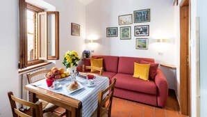 A cozy living room with a red sofa, a dining table with a checkered tablecloth, and a window with wooden shutters. - Corallo studio mezzanine (Spello)