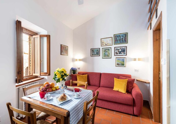 Un salon chaleureux avec un canapé rouge, une table à manger recouverte d’une nappe à carreaux et une fenêtre avec des volets en bois.