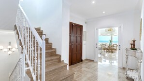 Una escalera con balaustres blancos que conduce a una puerta de madera, un comedor con vistas al mar y una consola blanca con un florero y velas.