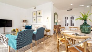 Una sala moderna con un sofá, dos sillas estampadas, una mesa de comedor con sillas, un televisor, cuadros enmarcados en la pared y un florero verde sobre la mesa.