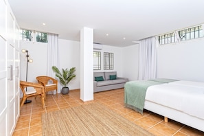 Una habitación de hotel moderna con cama, sofá, sillas de mimbre, planta en maceta y un ventanal con cortinas.