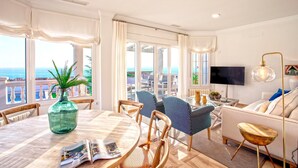 Una sala de estar moderna con un ventanal grande con vista al mar, un comedor con mesa y sillas, y un sofá con televisor.