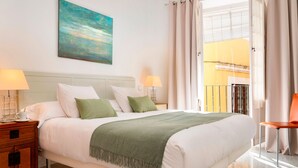A neatly made bed with a green throw, two bedside tables with lamps, a painting on the wall, and a balcony with curtains. - Genteel Home Pasaje Santa Cruz II (Seville)