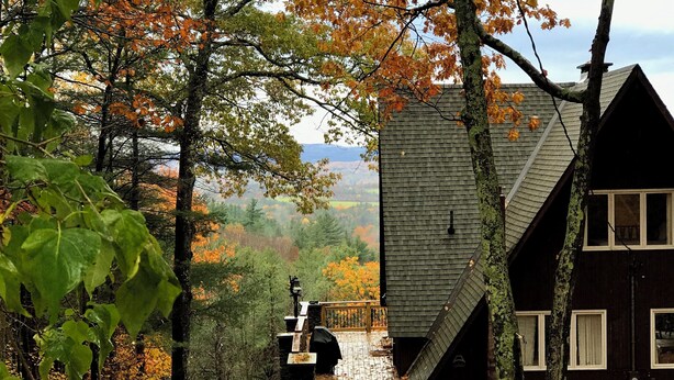 Eine Hütte mit steilem Dach, einer Holzterrasse und Blick auf ein bewaldetes Tal.