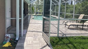 Une terrasse fermée par des baies vitrées, avec une piscine, du mobilier d’extérieur et une vue sur la verdure environnante.
