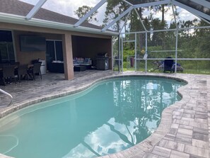 Une piscine avec un patio couvert, du mobilier d’extérieur et une vue sur les arbres environnants.