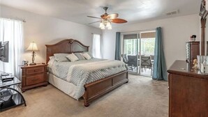 Une chambre avec un lit en bois, un ventilateur de plafond, un bureau avec une lampe et une vue sur l’extérieur grâce à une porte-fenêtre coulissante.