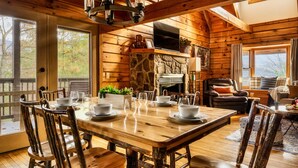 Interior de uma cabana de madeira com uma mesa de jantar posta para quatro pessoas, lareira e uma área de estar acolhedora.