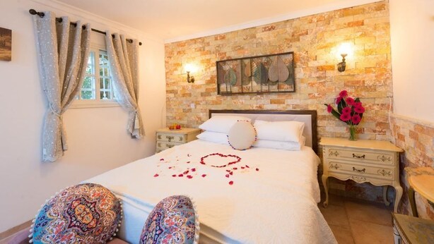 A bedroom with a brick wall, a bed with white linens, a nightstand, and a small table with a vase of flowers.