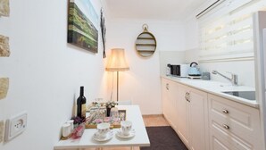A compact kitchen with white cabinets, a small dining table with floral tableware, a lamp, and a painting on the wall.
