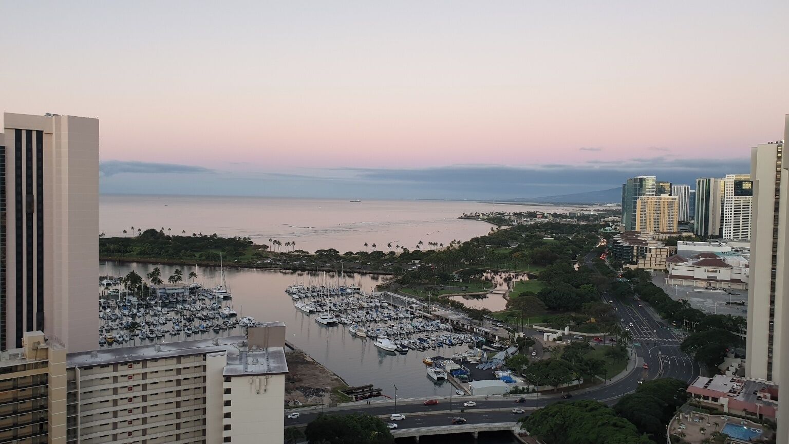 Un port de plaisance où sont amarrés de nombreux bateaux, entouré d’immeubles de grande hauteur et bordé par une route où circulent des véhicules.
