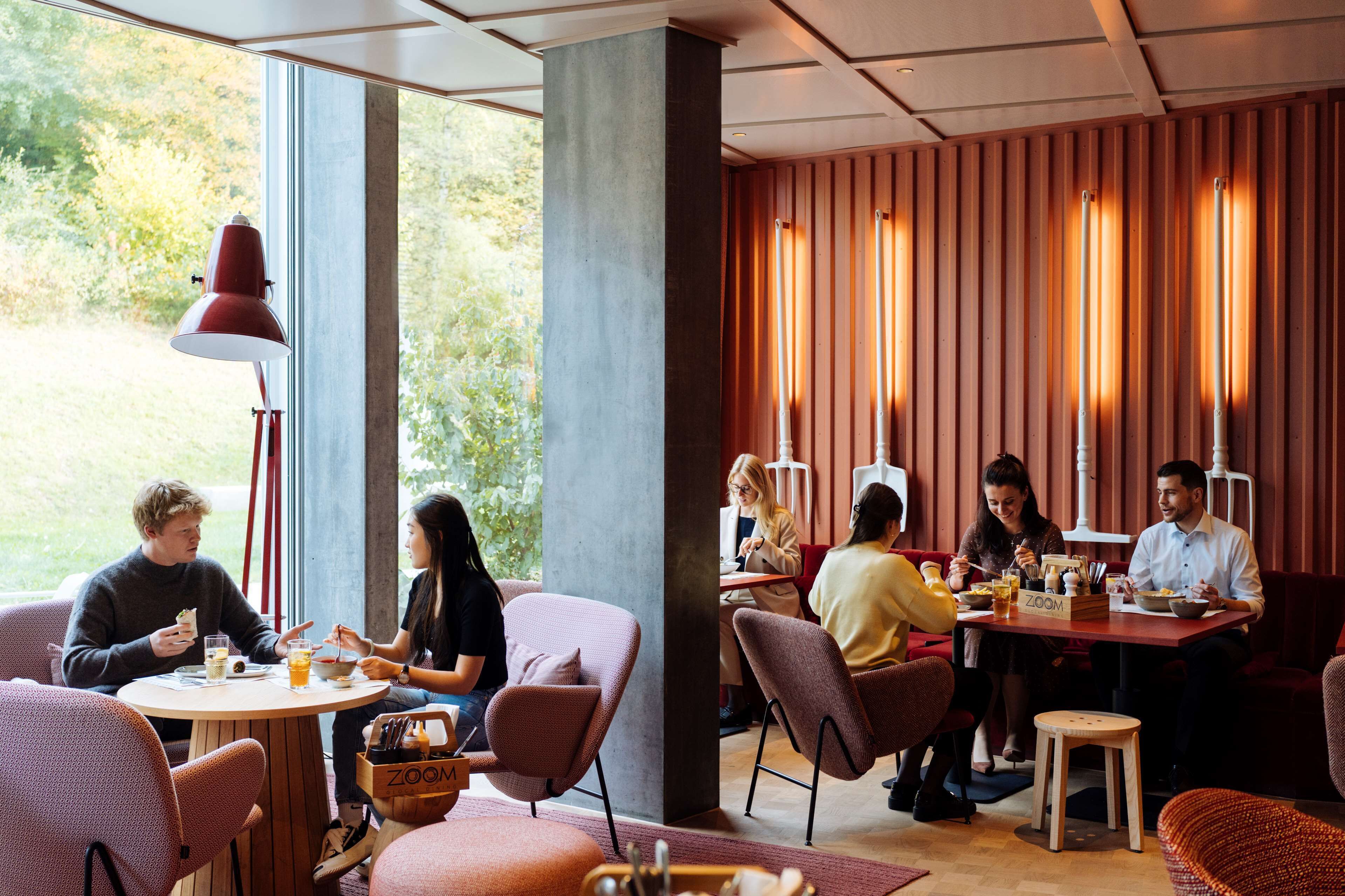Un espace de restauration moderne, doté de tables rondes, de sièges confortables et de grandes fenêtres offrant une vue sur la verdure.