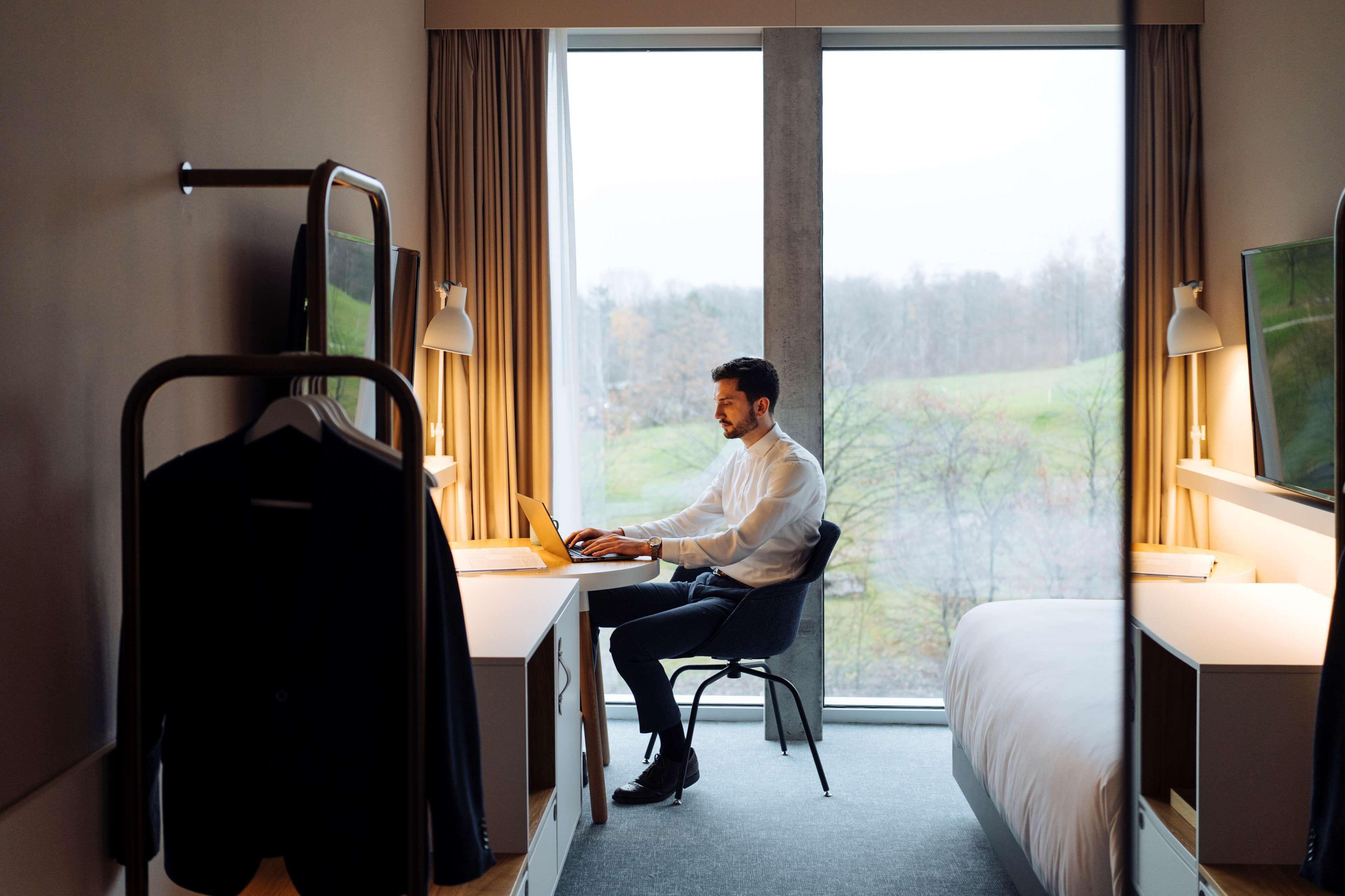 Una habitación de hotel con una cama, un escritorio con una computadora portátil, un perchero y un ventanal con vistas a árboles.