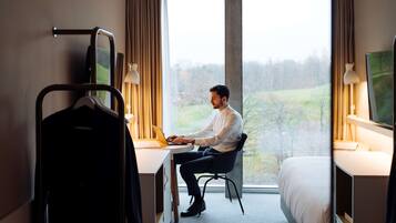 Una habitación de hotel con una cama, un escritorio con una computadora portátil, un perchero y un ventanal con vistas a árboles.