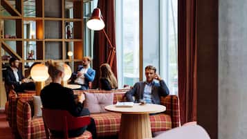 Un espace détente moderne avec des sièges rouges et à carreaux, une table ronde en bois et de grandes fenêtres avec des rideaux.