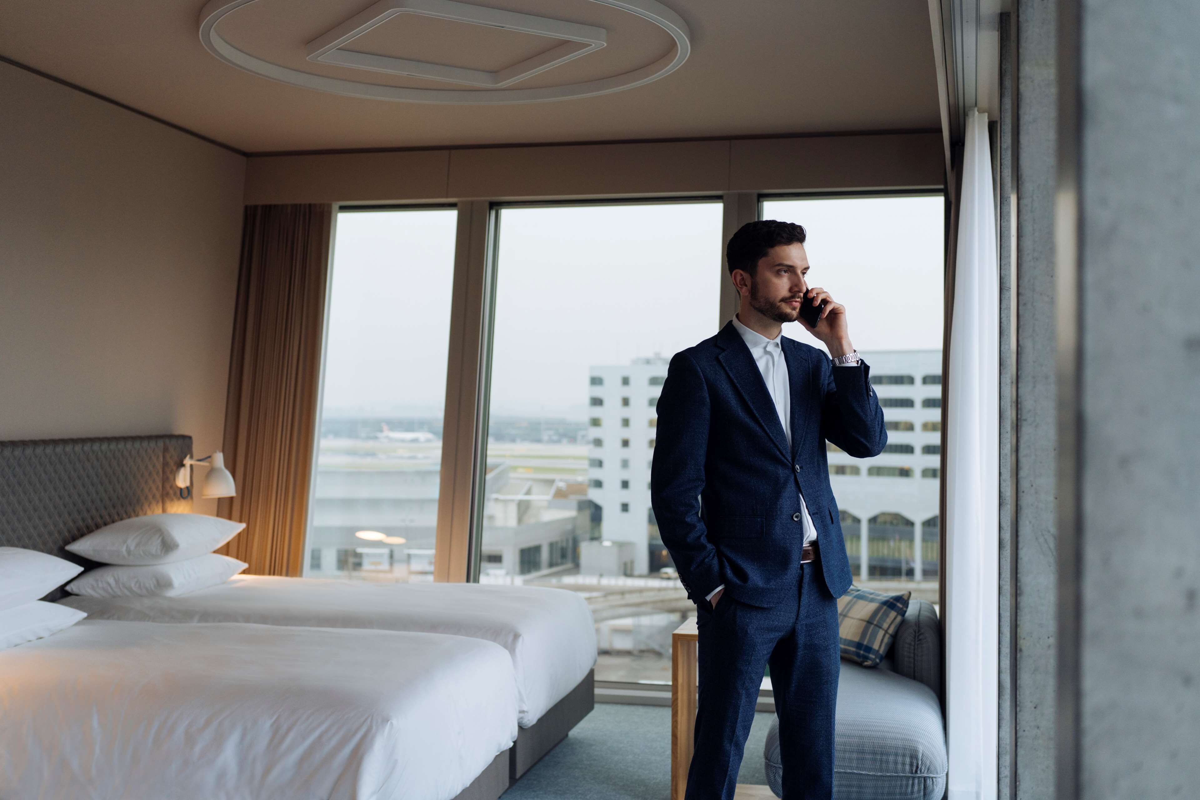 A hotel room with a large bed, a sofa, and a view of the cityscape through large windows.