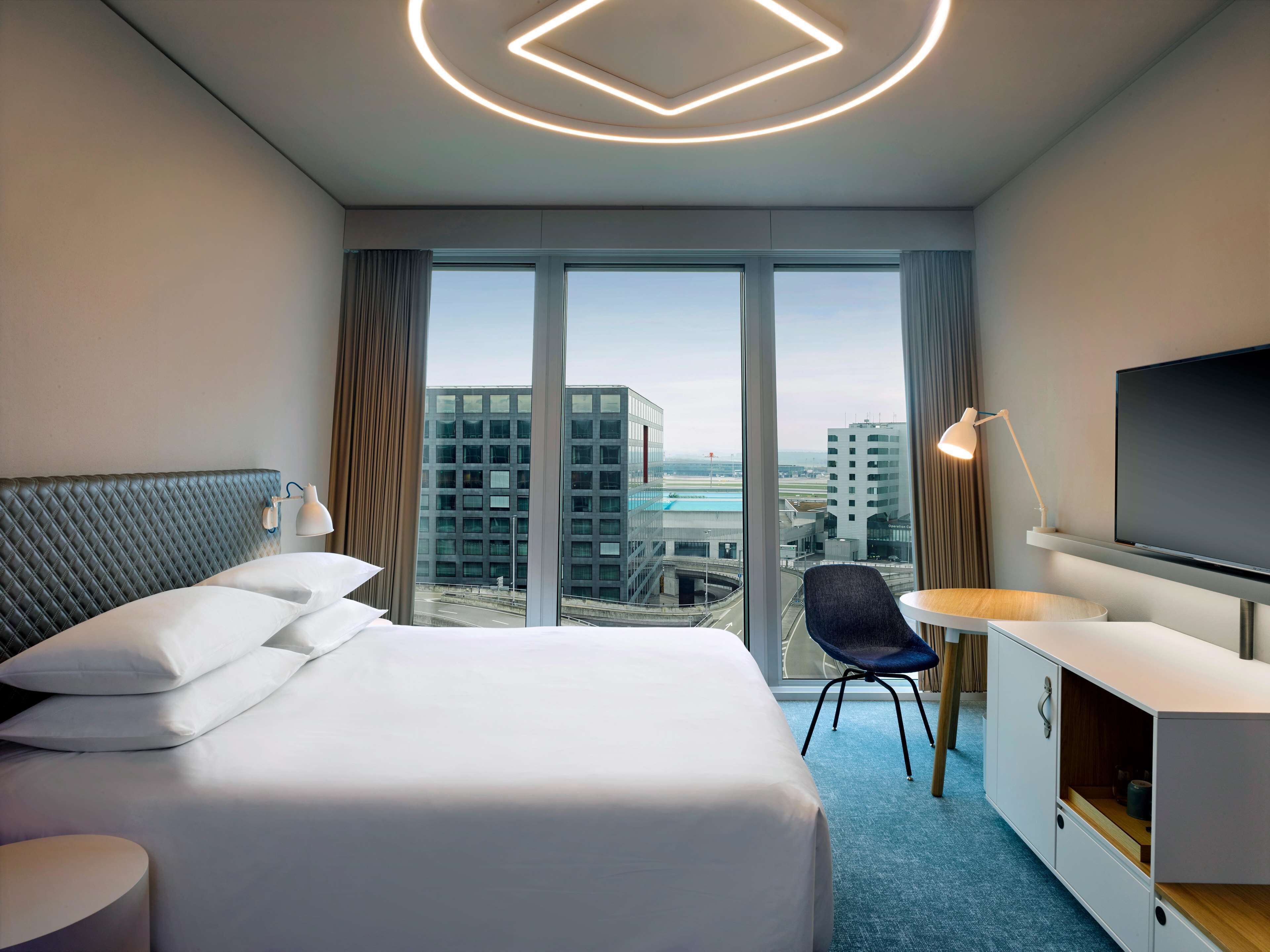 A hotel room with a large bed, a desk with a TV, and a view of the cityscape through large windows.