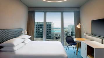 A hotel room with a large bed, a desk with a TV, and a view of the cityscape through large windows.