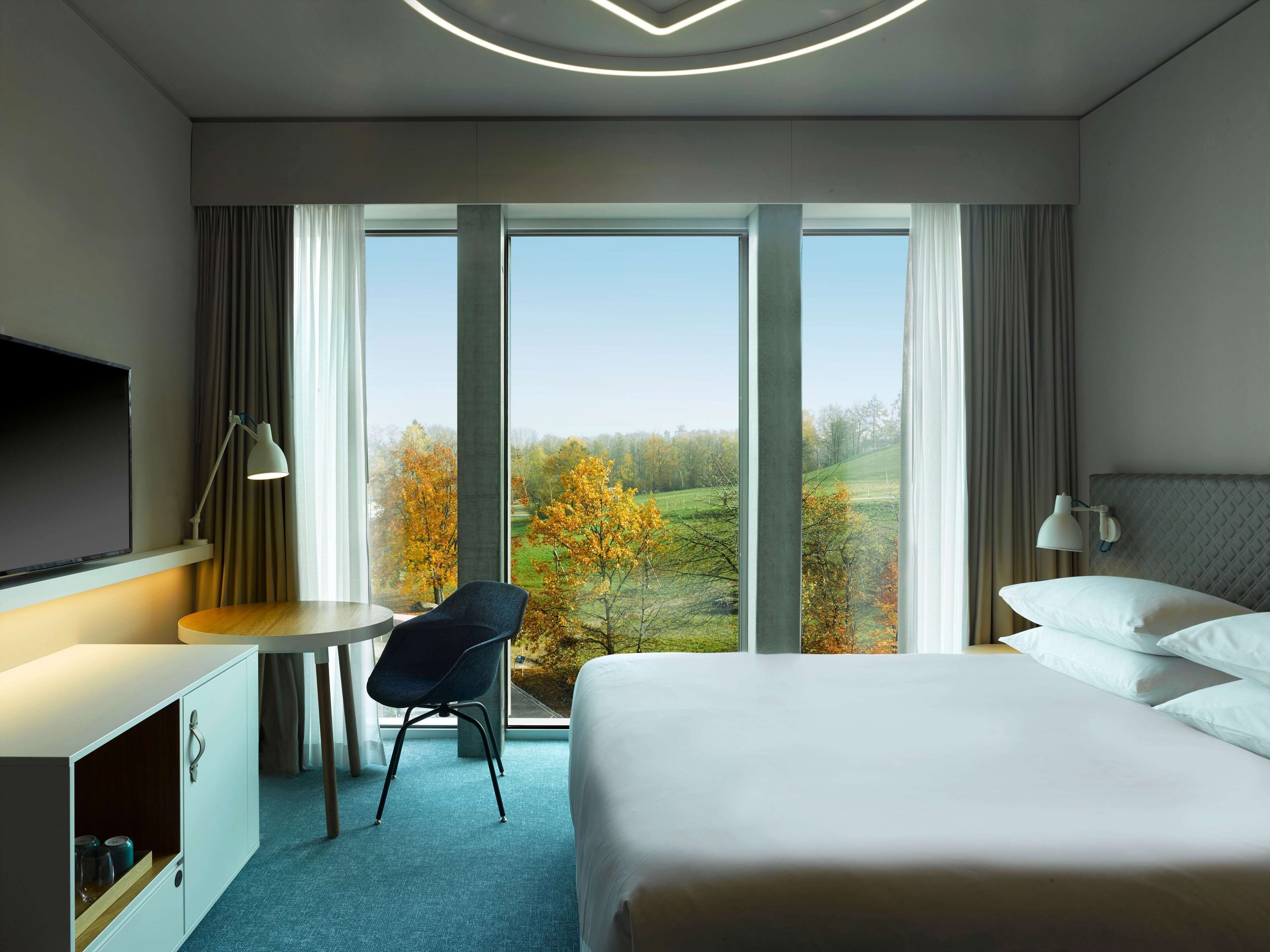 Een hotelkamer met een bed, een bureau, een stoel, een televisie en grote ramen met uitzicht op bomen.