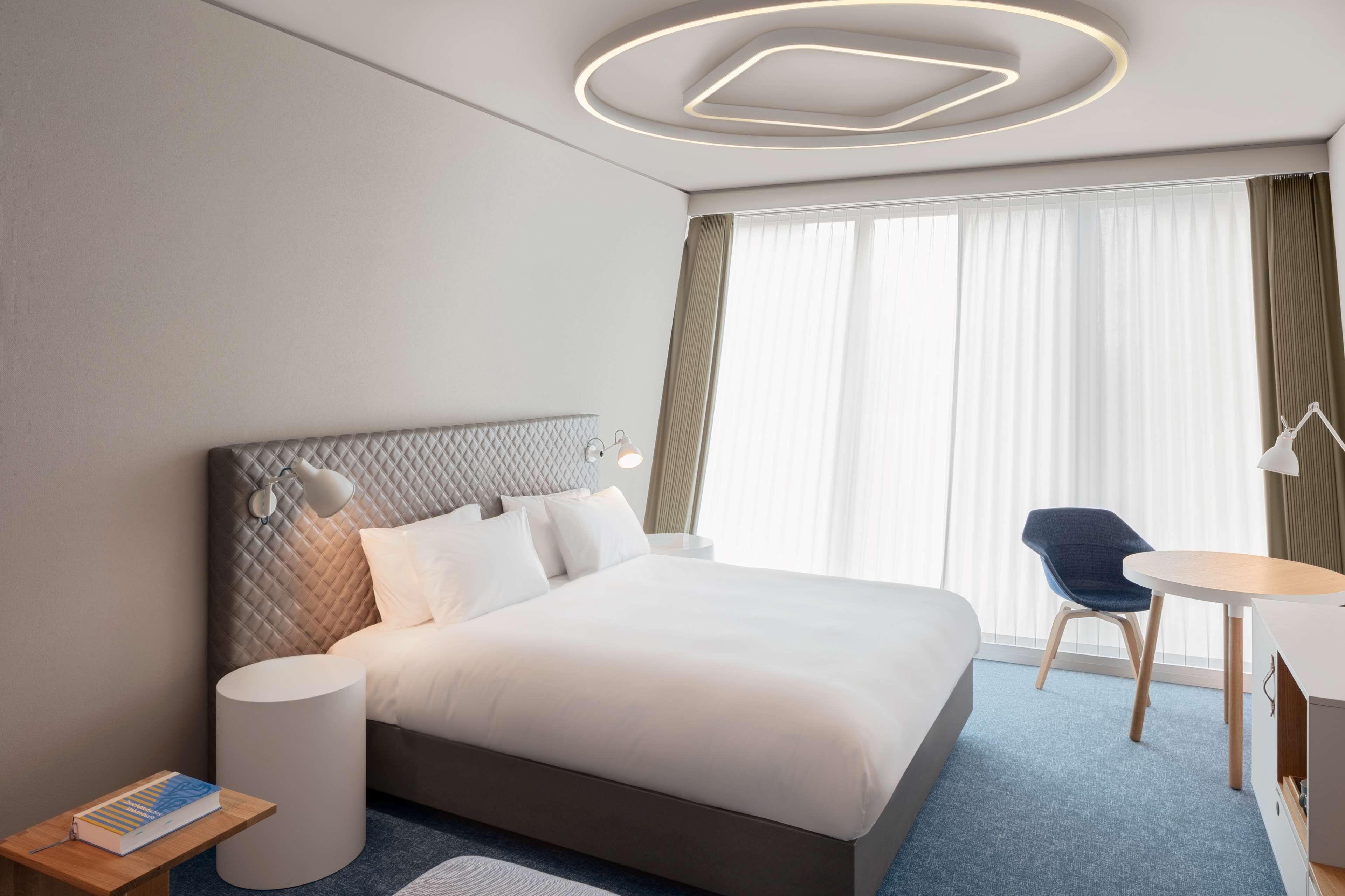 A modern hotel room with a large bed, a bedside table with a book, a round table with a chair, and a large window with curtains.