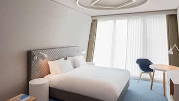 Una habitación de hotel moderna con una cama grande, una mesita de noche con un libro, una mesa redonda con una silla y un ventanal con cortinas.