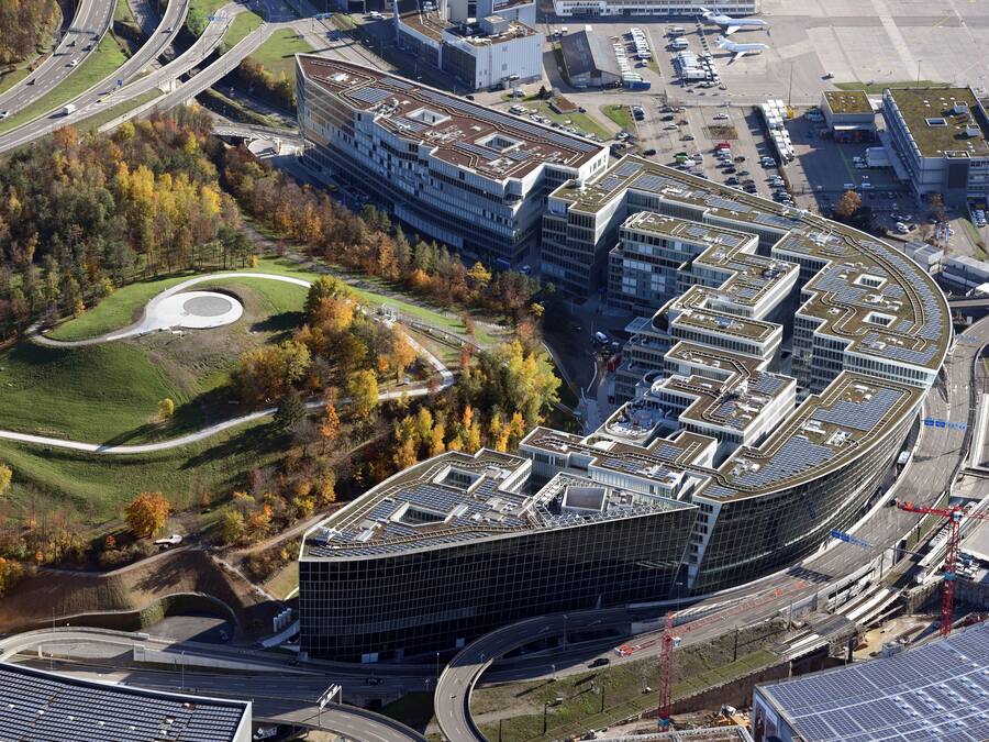 A modern building with a curved roof and multiple floors, surrounded by a landscaped area with trees and a circular park.