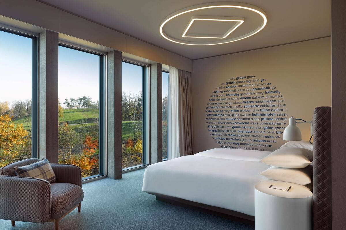 A modern hotel room with a large bed, a sofa, a bedside table with lamps, and a view of a green landscape through large windows.