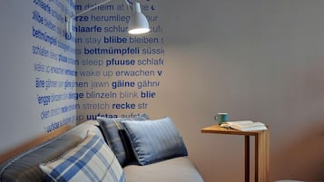 A cozy room with a sofa, pillows, a small table with a book and a cup, and a wall with text in a foreign language.