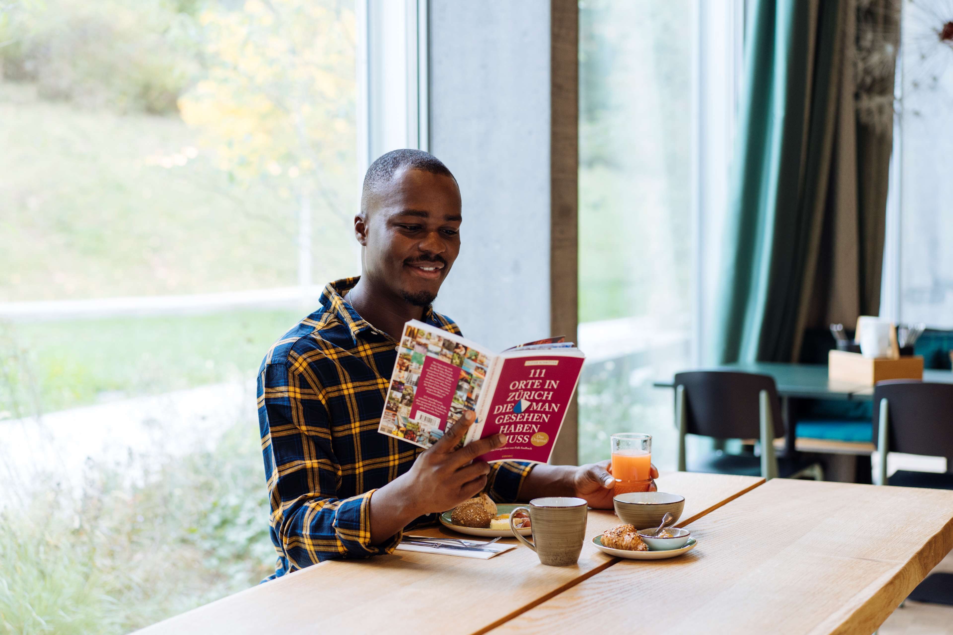 Una persona leyendo un libro en una mesa, con un vaso de jugo de naranja, un bol y un plato de comida.