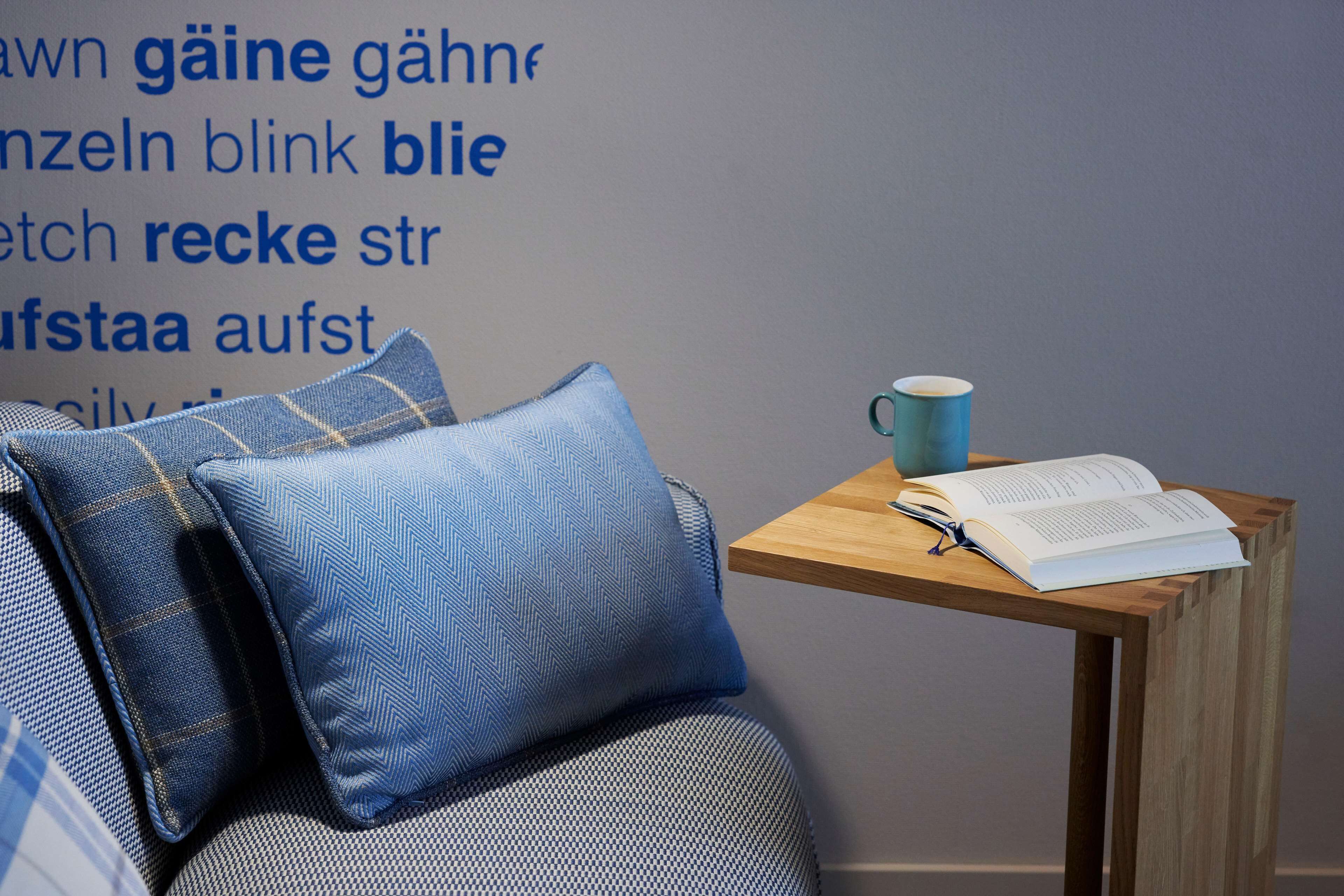Una habitación con una mesita de madera que sostiene un libro y una taza, un sofá azul con estampado, y una pared con texto en un idioma extranjero.