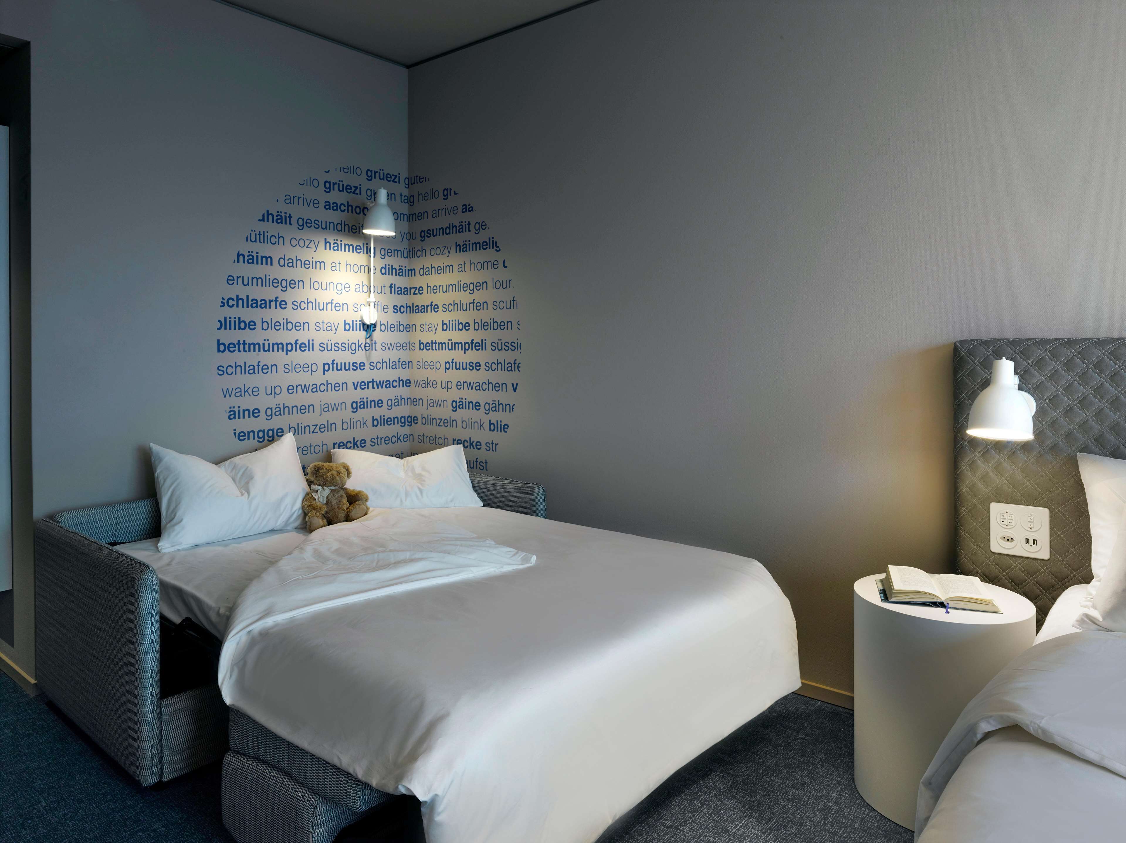A neatly made bed with white linens, a teddy bear, and a wall with text in a foreign language.