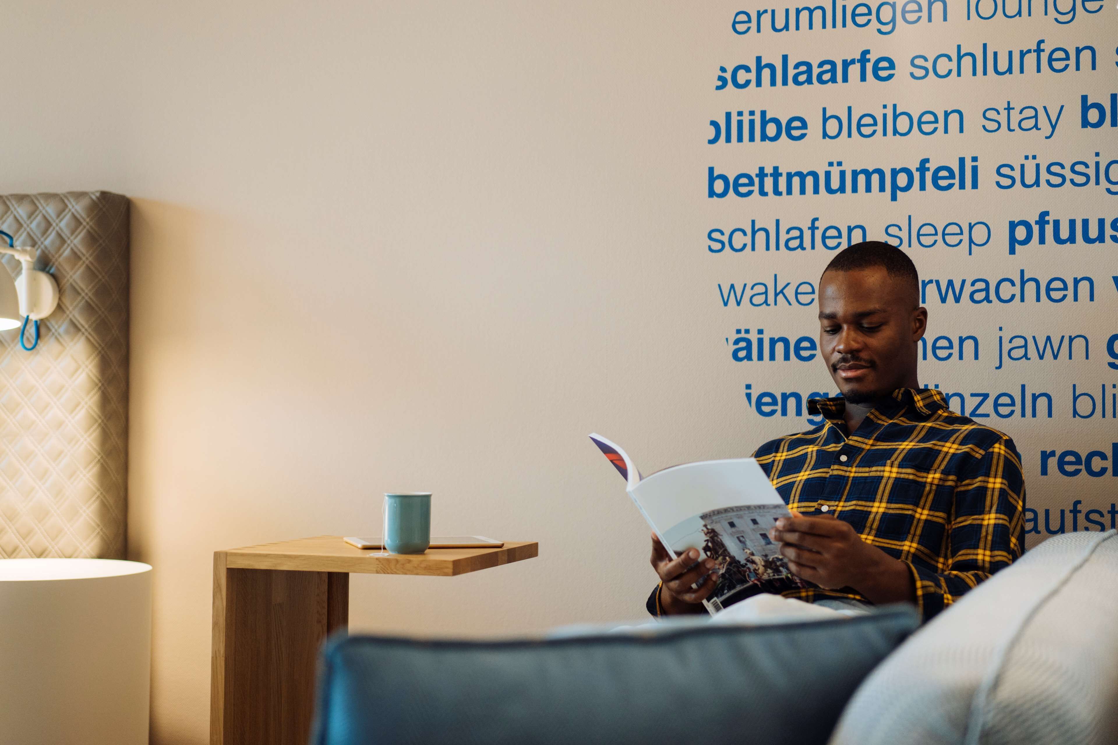A person reading a book in a room with a bed, a bedside table with a cup, and a wall with text in a foreign language.