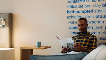 A person reading a book in a room with a bed, a bedside table with a cup, and a wall with text in a foreign language.