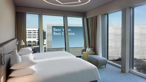 Una habitación de hotel moderna con una cama grande, una mesita de noche, una lámpara y vistas a la ciudad a través de amplios ventanales.