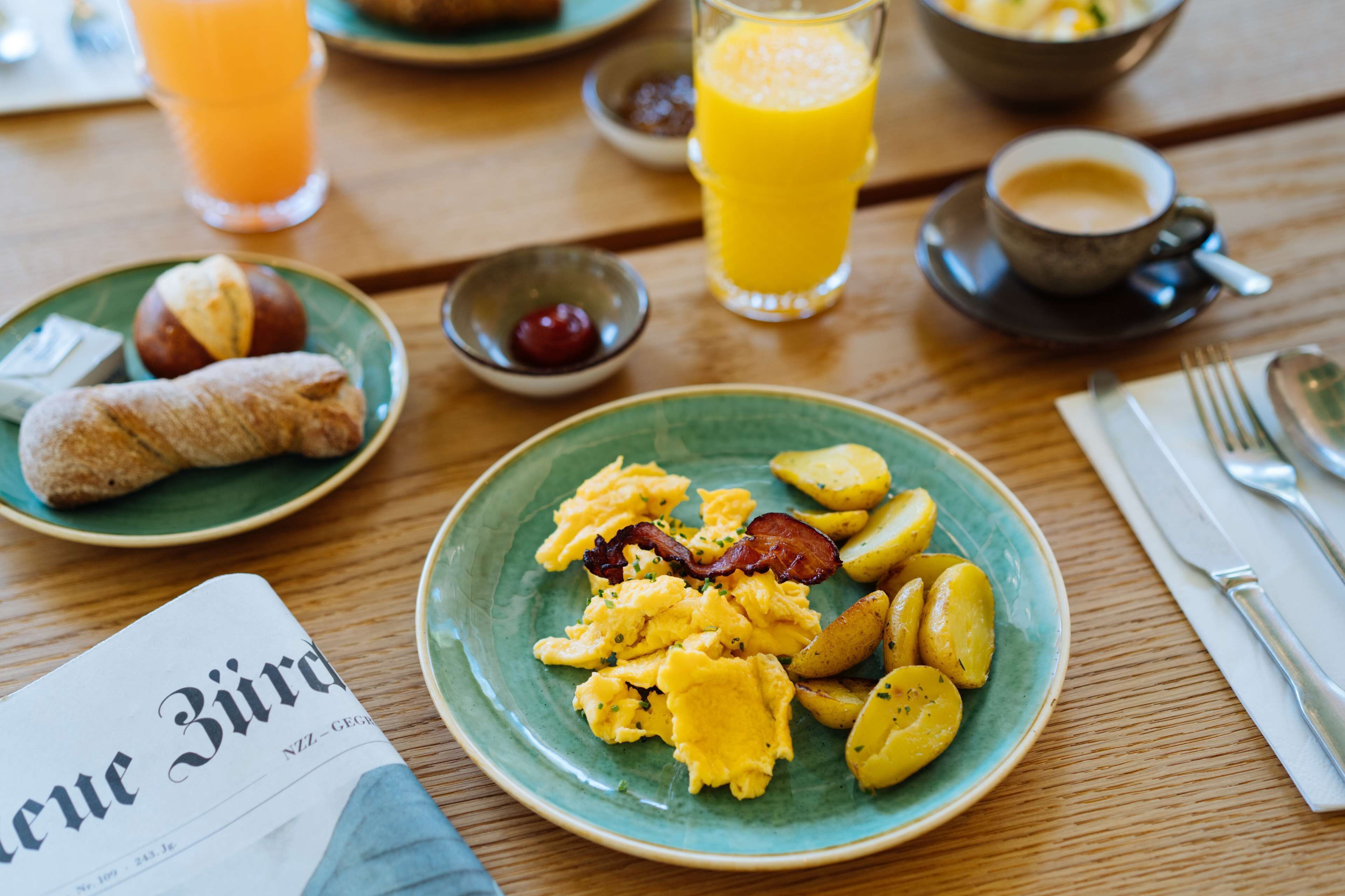 A breakfast setting with plates of scrambled eggs, bacon, and potatoes, a glass of orange juice, and a cup of coffee on a wooden table.