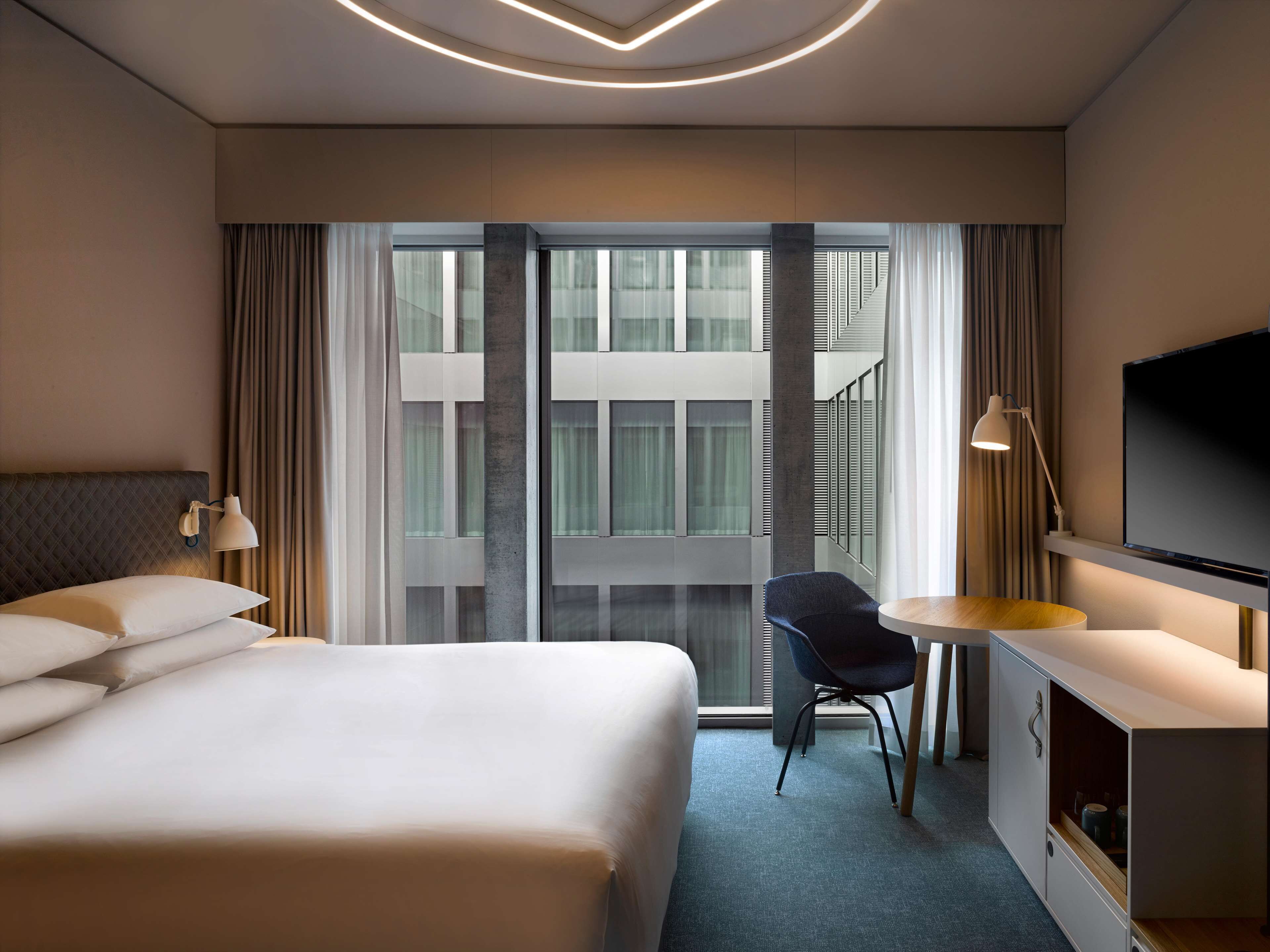 A modern hotel room with a large bed, a desk with a chair, a TV, and a view of the building's exterior through large windows.
