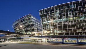 Un edificio moderno con amplios ventanales y una estación de tren al frente.