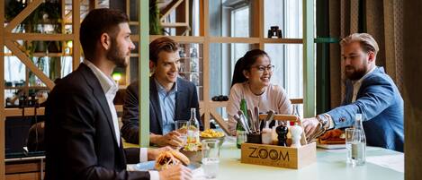 A group of people seated around a table with food and drinks, in a room with wooden partitions and large windows.