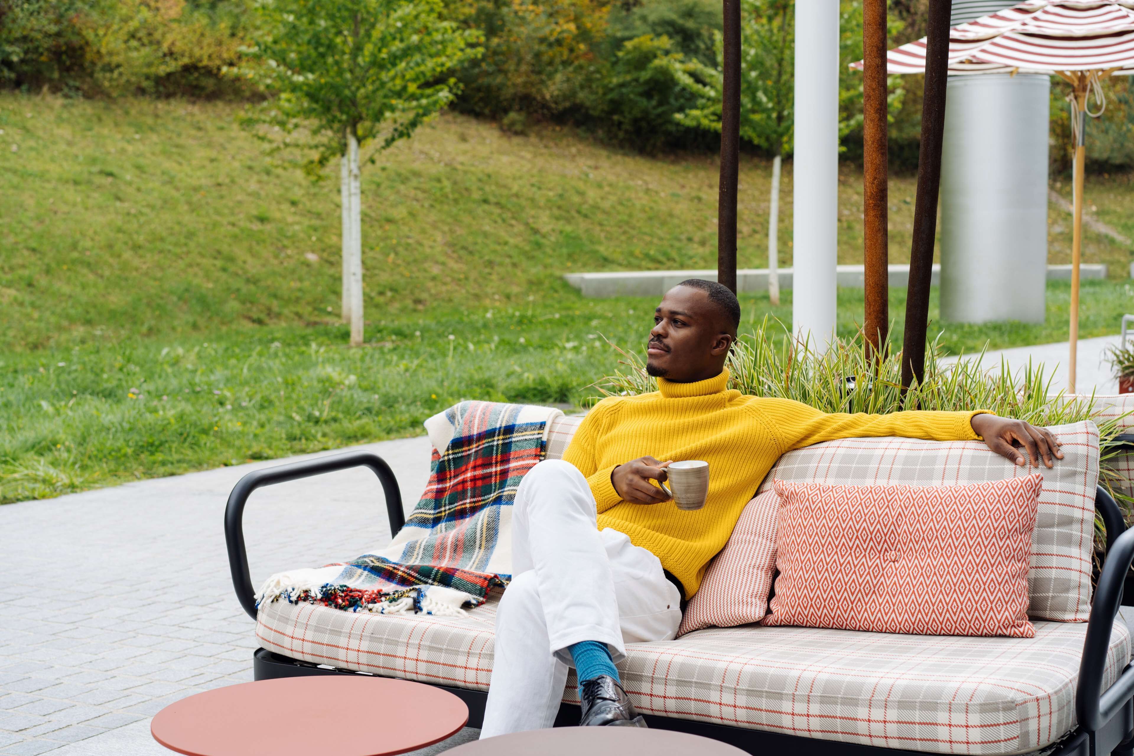 A person sitting on a plaid sofa with a cup, outdoors with grass and trees in the background.