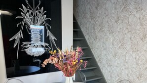 Una mesa redonda blanca con un florero blanco de flores secas, rodeada de sillas de plástico transparente, en una habitación con papel tapiz estampado y una lámpara moderna.