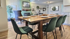 Un coin repas avec une table en bois dressée pour quatre personnes, des chaises à l’assise verte et, au fond, une cuisine moderne.