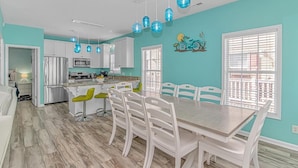 A modern kitchen with white cabinets, a dining area with a wooden table and white chairs, and blue pendant lights. - COCONUT TREEHOUSE, 5 bed/3.5 bath, 1 minute walk to beach, Holiday Inn section (Myrtle Beach)