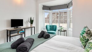 Ein modernes Hotelzimmer mit einem Bett, einem Fernseher, einem kleinen Tisch mit einer Pflanze, einem petrolfarbenen Sessel und Blick auf die Stadt durch große Fenster.
