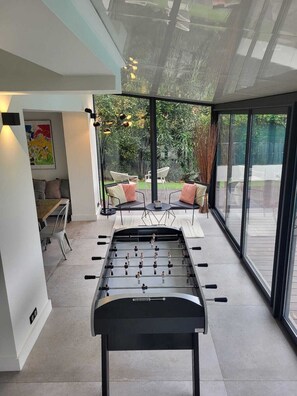 A modern interior with a foosball table, a dining area, and large windows offering a view of the outdoors.
