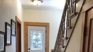 Un couloir doté d’un plancher en bois, d’un paillasson noir à l’entrée, d’une porte blanche ornée d’un vitrail et d’un escalier avec une rampe en bois.