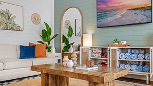 Ein Wohnzimmer mit einem Holztisch, einem weißen Sofa mit bunten Kissen, einem großen Spiegel und einem Fernseher, der eine Strandansicht zeigt.
