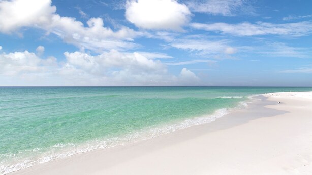Ein Strand mit klarem türkisblauem Wasser und sandigem Ufer unter einem teilweise bewölkten Himmel.