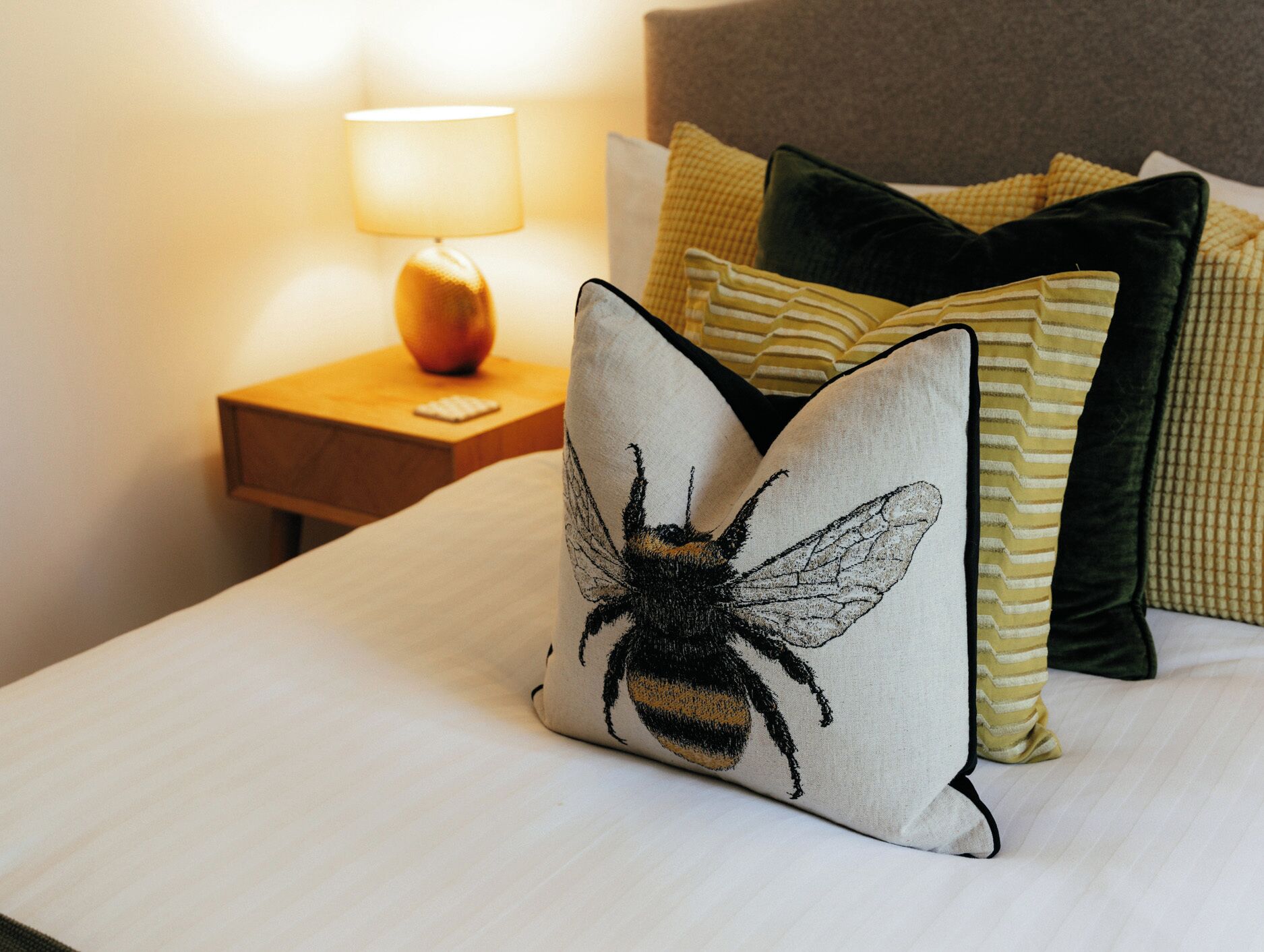 A neatly made bed with a pillow featuring a bee design, a lamp on a bedside table, and striped pillows.